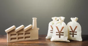 Photo of bags of Japanese yen next to a wooden representation of house/factory.