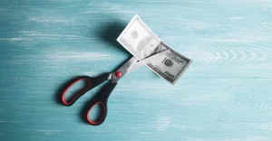 A pair of scissors cutting a dollar bill on a blue wooden table.