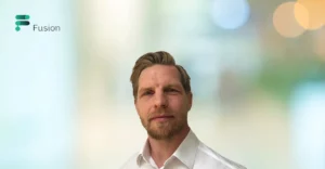 A man in a white shirt is standing in front of a blurry background.