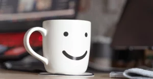 A mug with a smiley face on it sits on a desk.