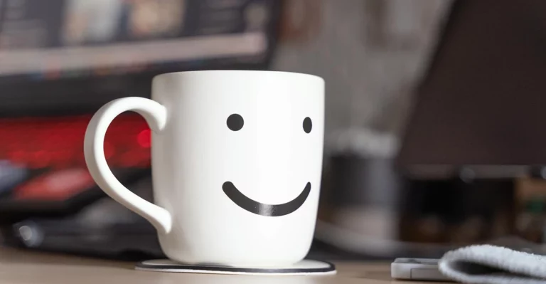 A mug with a smiley face on it sits on a desk.