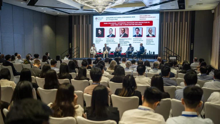 Panel discussion on stage at a conference, five speakers seated with a large screen behind them displaying bios and a red banner.