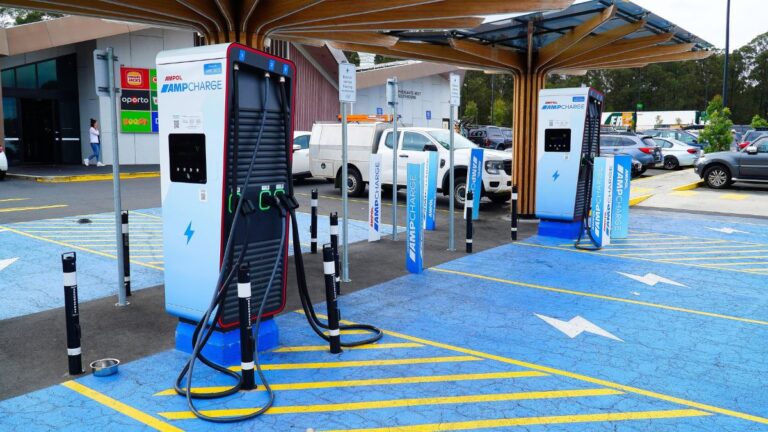 Electric vehicle charging station area with multiple AmpCharge chargers under a wooden canopy, blue-marked parking spaces, and cars in the background.