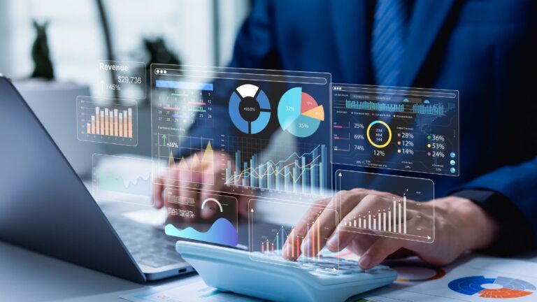 Businessperson analyzing financial data with translucent holographic dashboards and charts over a laptop and calculator’s keyboard, in a bright office.