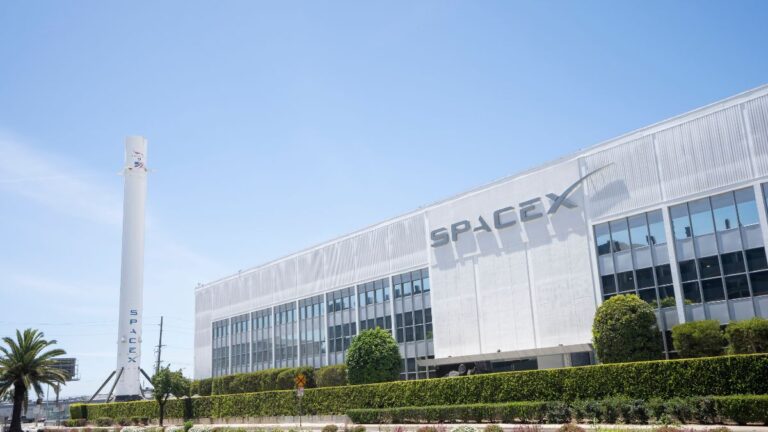 SpaceX facility with a tall rocket launch tower beside a modern building on a sunny day