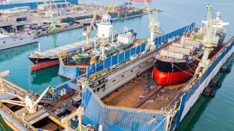 Aerial view of a busy shipyard with several large cargo ships in dry docks, cranes towering above, and turquoise water surrounding the docks.