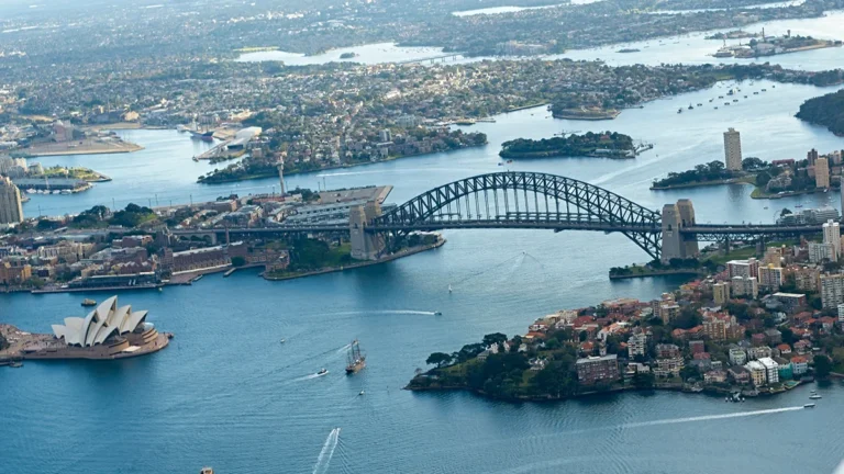 Aerial view of Sydney Harbour showing the Opera House and the Harbour Bridge with surrounding city and water traffic. The Opera House is on the left, the Bridge spans the harbor.