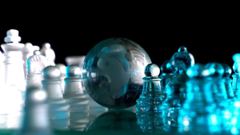 Close-up of a blue-tinted glass chess set on a reflective surface, with a translucent globe in the center against a dark background.