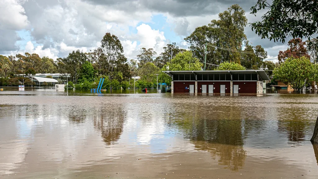 Flooded park scene with brown water surrounding a small red-brick building, trees, and blue playground structures, under a cloudy sky with reflections on the water.