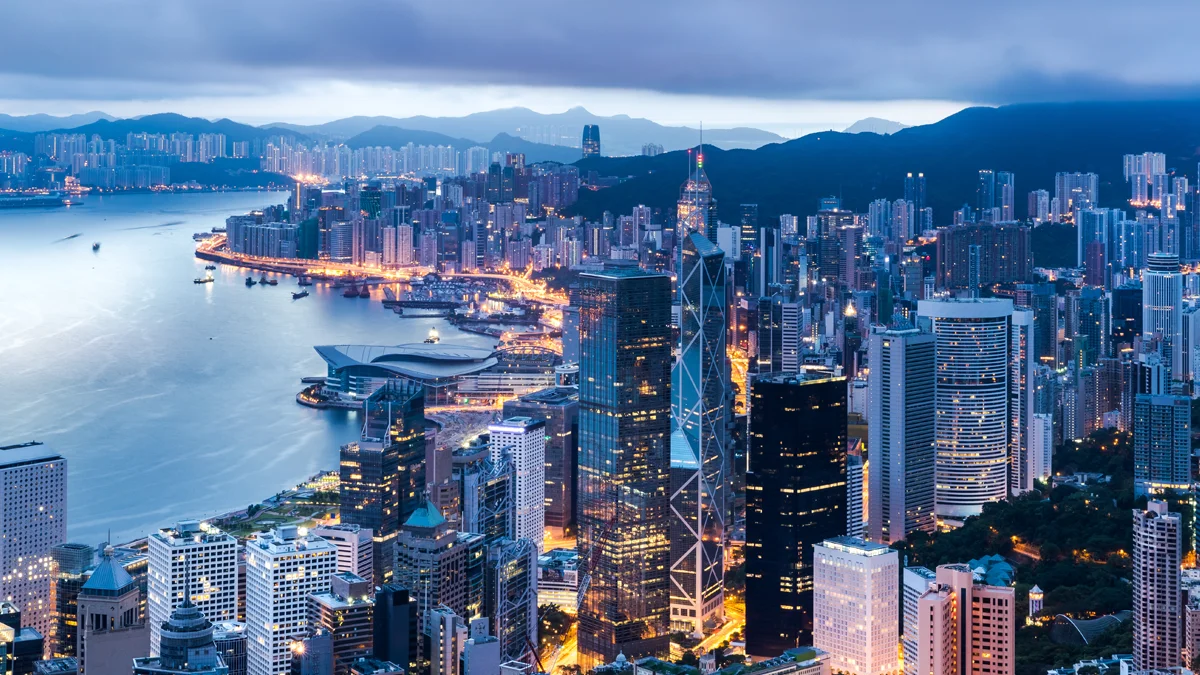 Evening city skyline of Hong Kong with tall skyscrapers along Victoria Harbour and mountains in the background.
