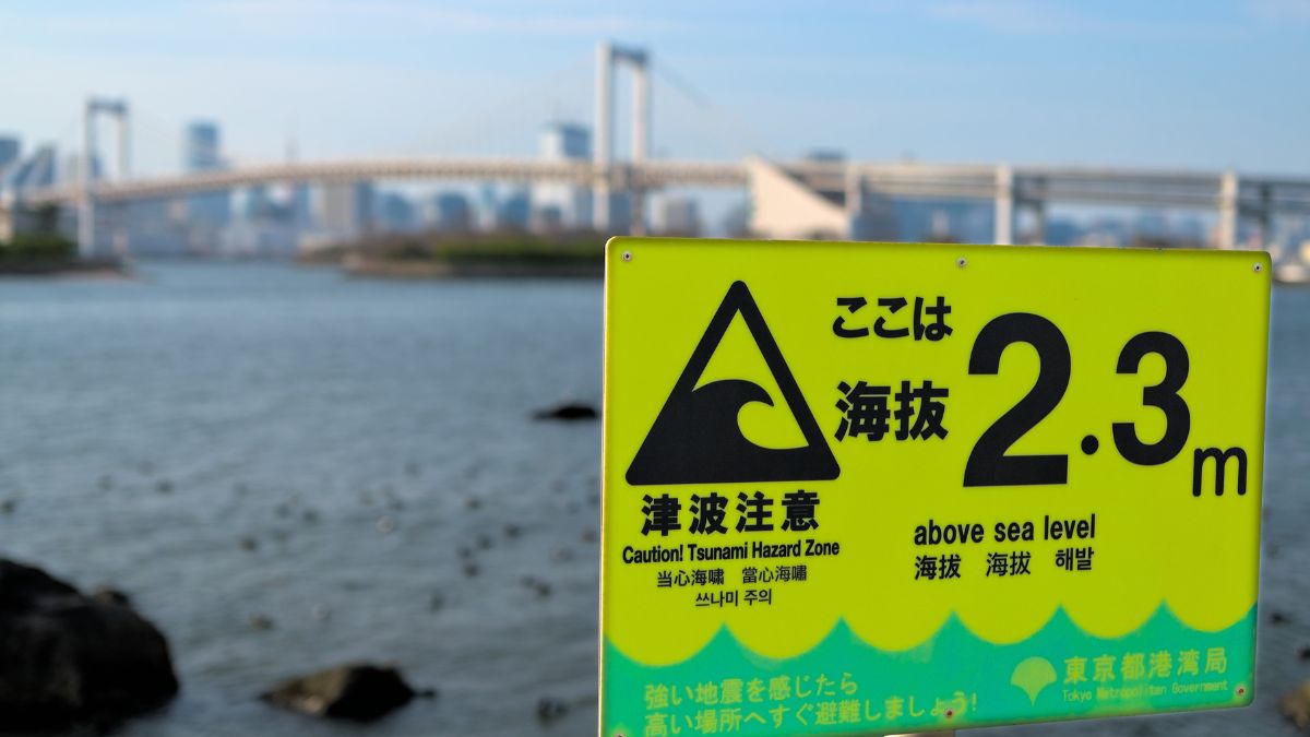 Bright yellow tsunami hazard sign by the water reading 'Caution! Tsunami Hazard Zone' with '2.3m above sea level' and a bridge in background.