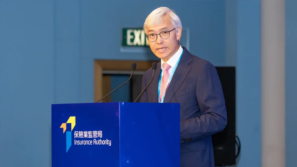 Man in a navy suit and pink tie giving a speech at a blue podium labeled Insurance Authority.
