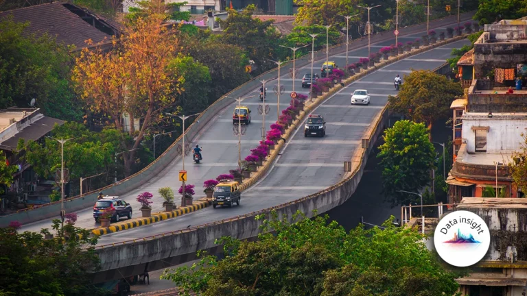 Rising highway with a central flower-lined divider, cars and motorbikes moving along, surrounded by trees and buildings; a circular Data Insight logo is in the bottom-right corner.