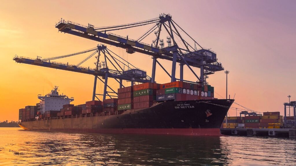 Container ship at a busy port during sunset, with towering gantry cranes and stacked shipping containers on deck.