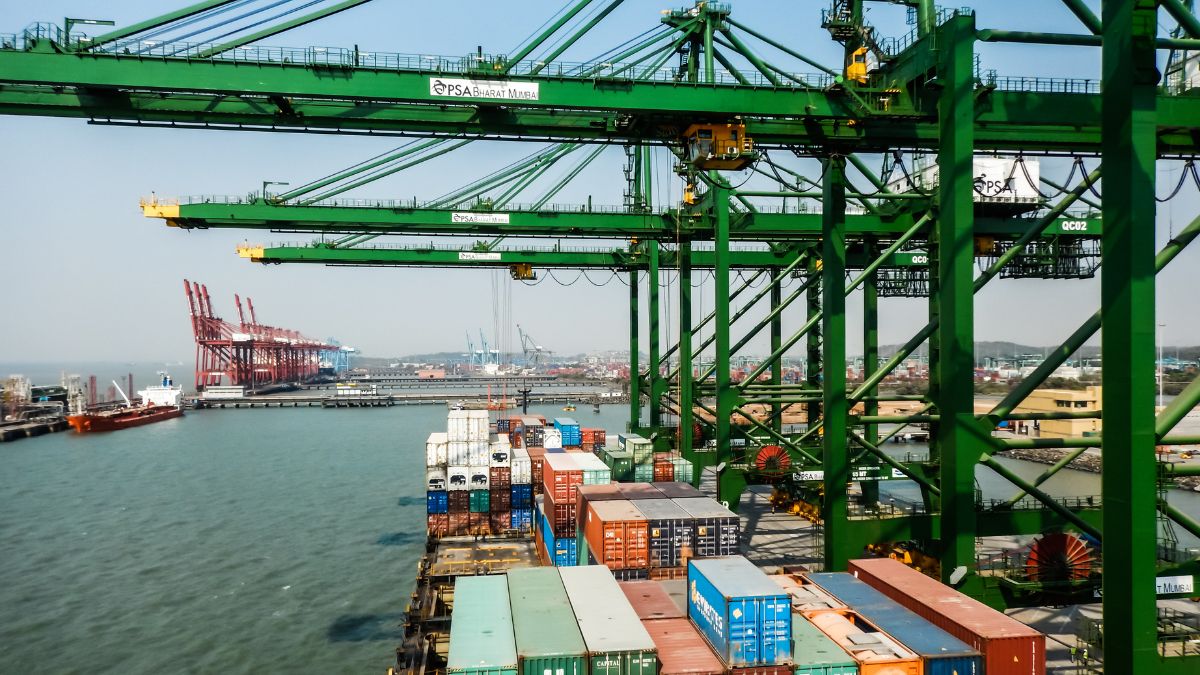 Green container cranes loom over a docked port with stacked cargo containers by the water.