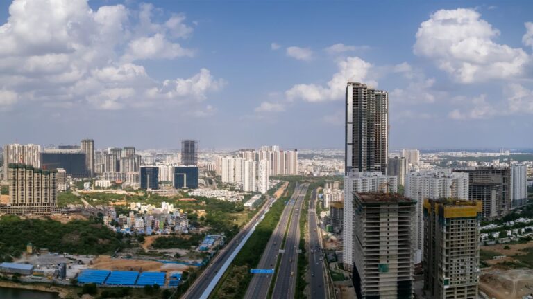 A sprawling cityscape with a wide, multi-lane highway cutting through high-rise buildings under construction under a blue sky.