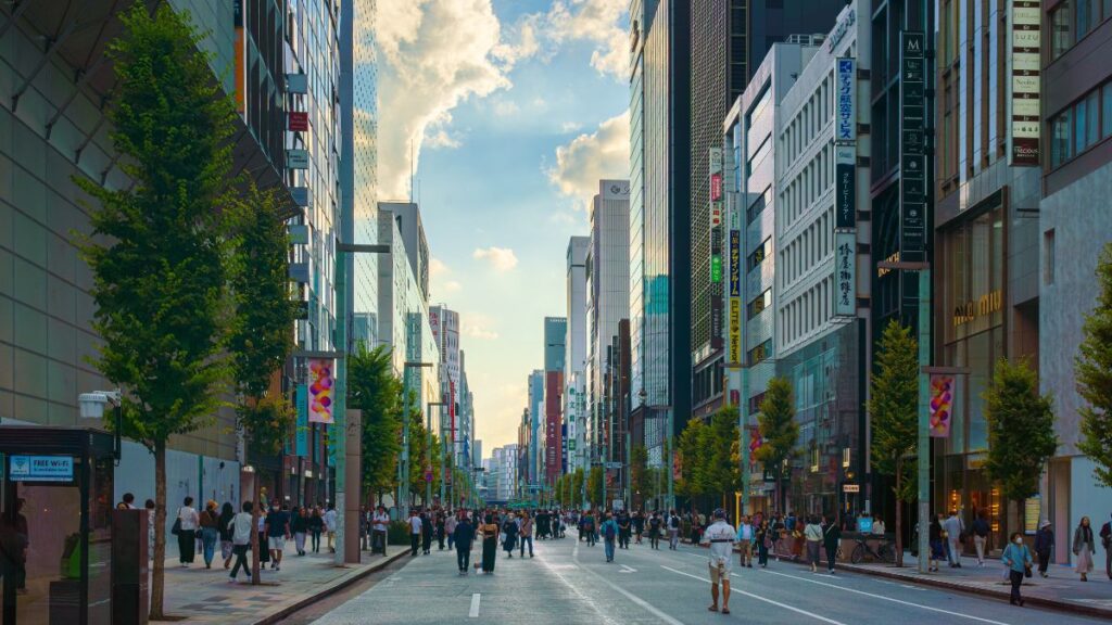 Busy city street lined with tall glass buildings and trees, pedestrians walking on both sides under a blue sky and scattered clouds.
