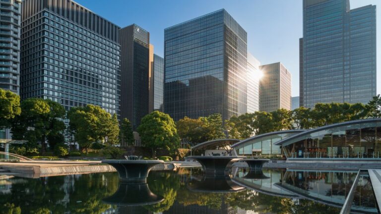 Modern city skyline with glass skyscrapers, a reflecting pool, and green trees in the foreground.
