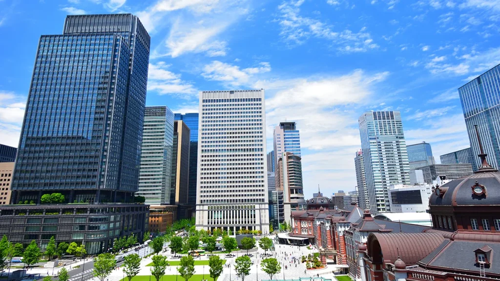 Wide city skyline with tall glass skyscrapers, blue sky, and a tree-lined plaza in the foreground. Central modern buildings flank a historic red-roofed structure to the right.