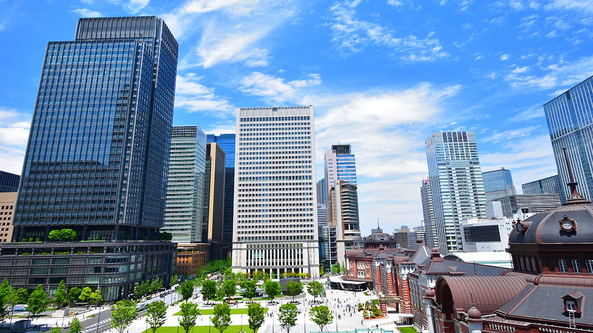 Wide city skyline with tall glass skyscrapers, blue sky, and a tree-lined plaza in the foreground. Central modern buildings flank a historic red-roofed structure to the right.