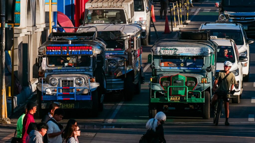 Colorful Philippine jeepneys and other vehicles line a busy city street, with pedestrians and a cyclist crossing nearby.