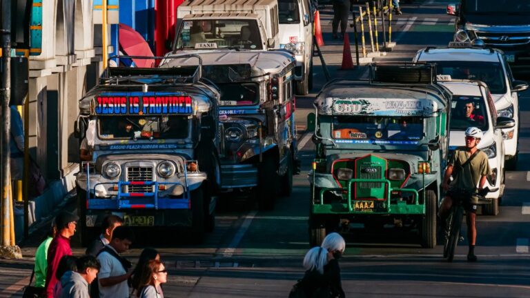 Colorful Philippine jeepneys and other vehicles line a busy city street, with pedestrians and a cyclist crossing nearby.