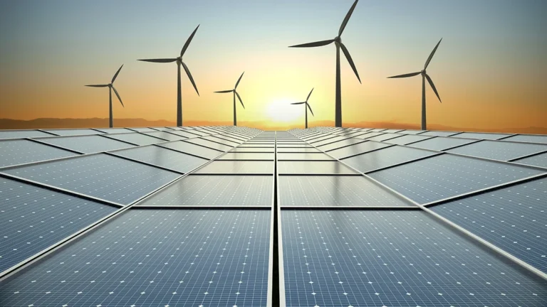 Solar panels stretch toward the sunset with wind turbines silhouetted on the horizon, depicting renewable energy.