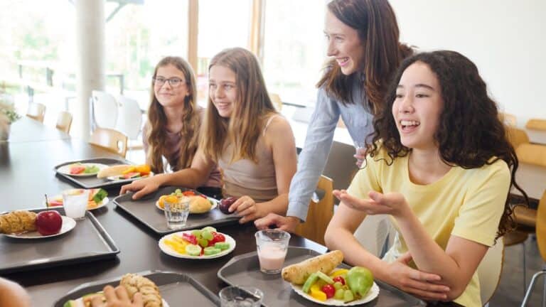 Four girls and a supervising adult at a cafeteria table, chatting and eating lunch items like fruit, bread, and vegetables.