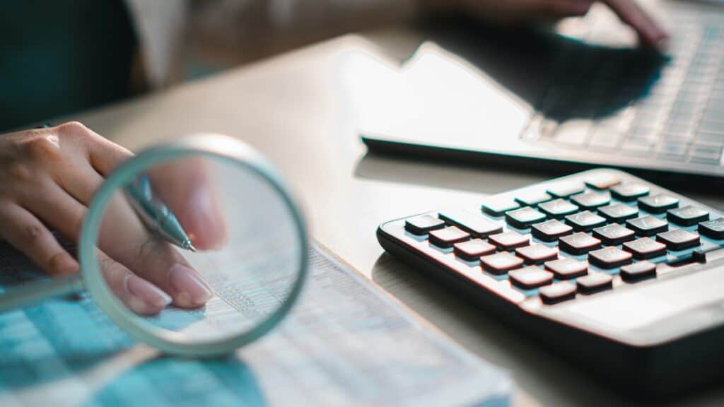 Person analyzes data on a desk, using a pen under a magnifying glass with a calculator and laptop nearby
