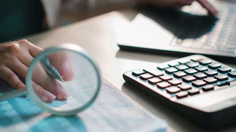 Person analyzes data on a desk, using a pen under a magnifying glass with a calculator and laptop nearby