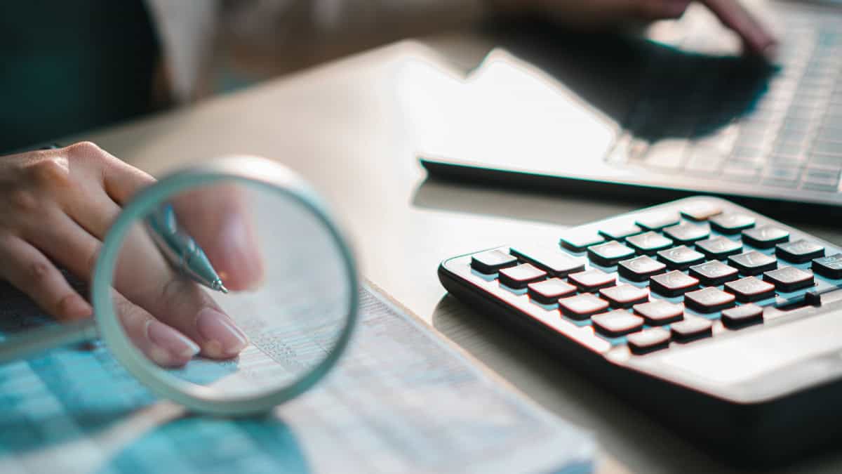 Person analyzes data on a desk, using a pen under a magnifying glass with a calculator and laptop nearby
