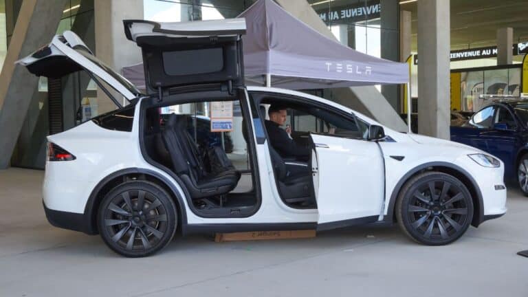 White Tesla Model X with falcon wing doors raised, a man sits in the driver’s seat inside a showroom.