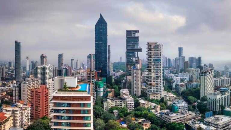 Panoramic city skyline with tall glass skyscraper and apartment blocks, a rooftop pool in the foreground, and an overcast sky.