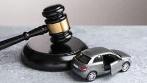 Gavel beside a silver toy car with an open door on a gray surface, suggesting auto law or car accident legal matters.