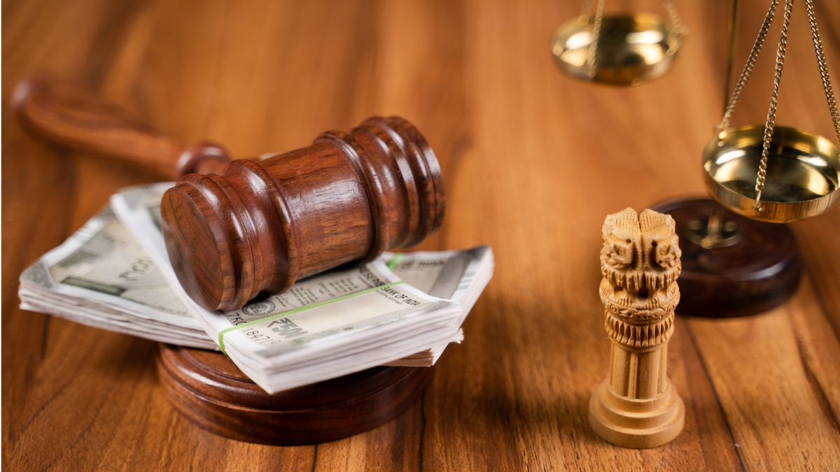 Judge's gavel resting on a stack of cash with a scales of justice in the background, on a wooden table.
