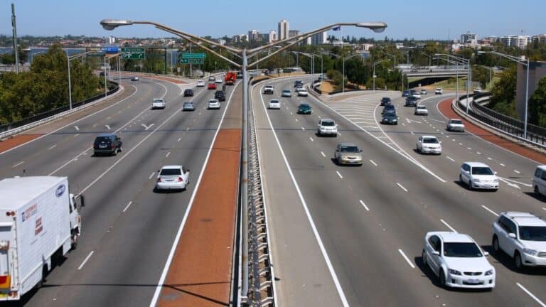 Multi-lane highway with a central barrier and cars traveling in both directions, urban skyline in the distance under a clear blue sky.