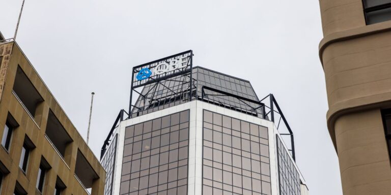 Modern glass office building seen from below with a blue company logo sign atop.