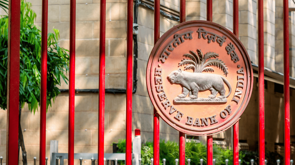 Seal of the Reserve Bank of India on a red gate with a tiger and palm tree emblem in relief behind bars outside a stone building.