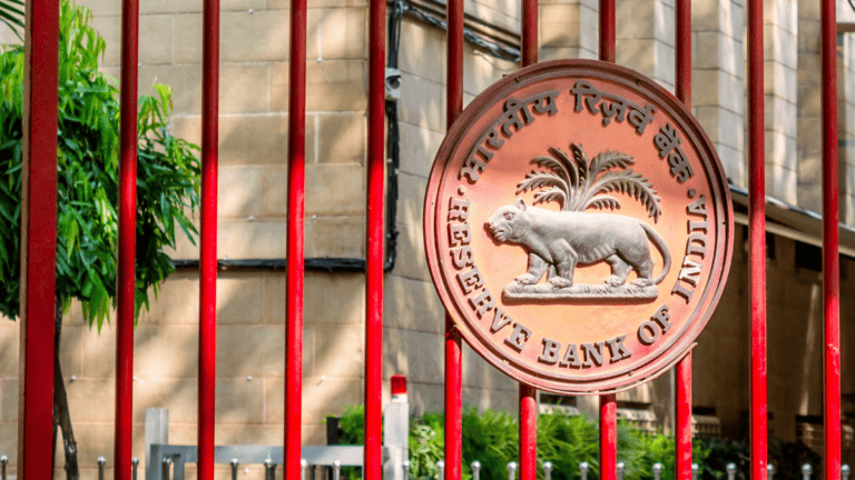 Seal of the Reserve Bank of India on a red gate with a tiger and palm tree emblem in relief behind bars outside a stone building.