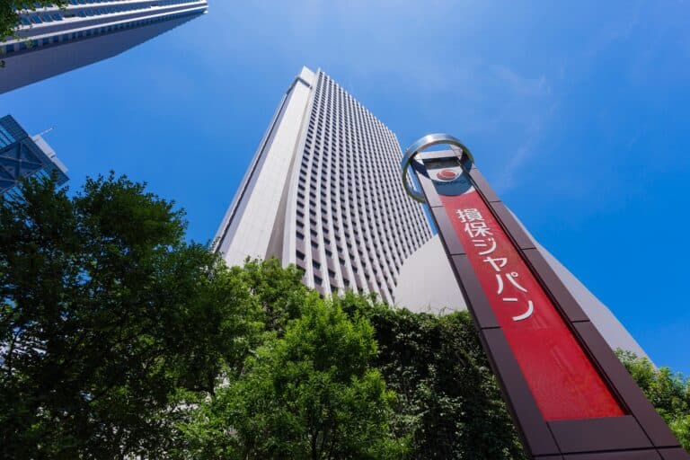 Low-angle view of tall white skyscrapers against a clear blue sky, with a red Sompo banner and green trees in the foreground.