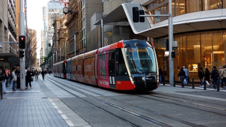 Red modern tram glides along tracks on a busy city street, with pedestrians on either sidewalk and tall office buildings nearby.
