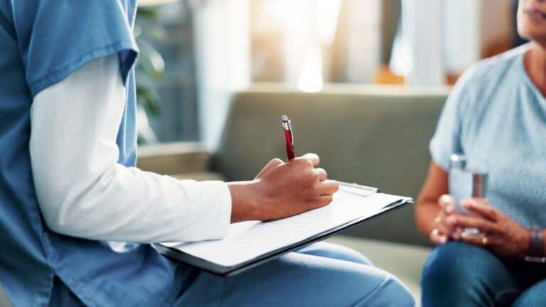 Healthcare professional in blue scrubs writes on a clipboard during a consultation with a patient seated nearby holding a bottle of water