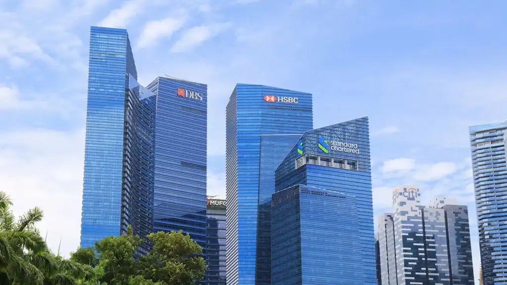 Blue glass skyscrapers in a financial district with bank logos DBS, HSBC and Standard Chartered visible on the towers, against a blue sky with palm trees in the foreground