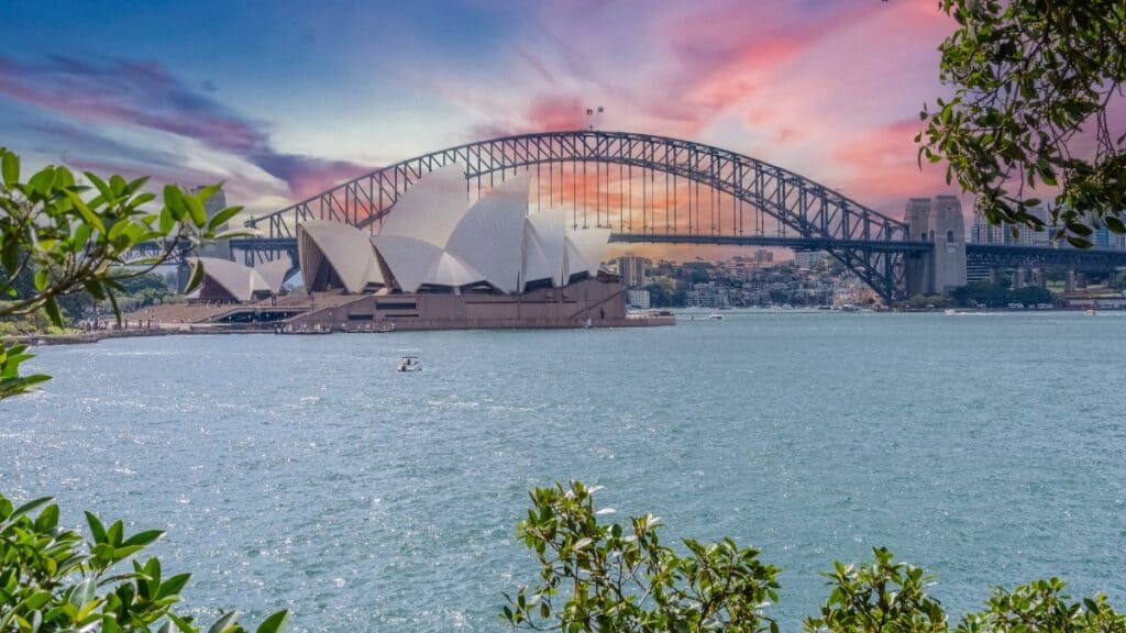 Sunset view of Sydney Harbour: the Opera House with the Harbour Bridge in the background over calm water, framed by green foliage on the sides.