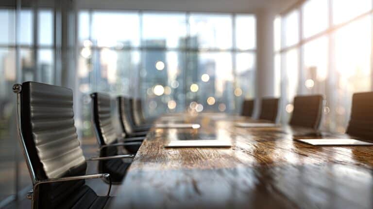 Empty modern conference room with a long wooden table and black leather chairs, large windows with blurred city lights in the background.