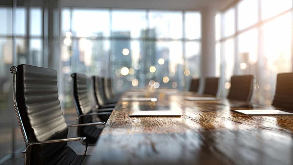 Empty modern conference room with a long wooden table and black leather chairs, large windows with blurred city lights in the background.