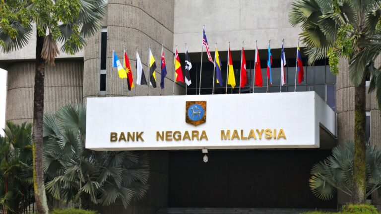 Front of Bank Negara Malaysia building with a row of international flags and palm trees on either side.
