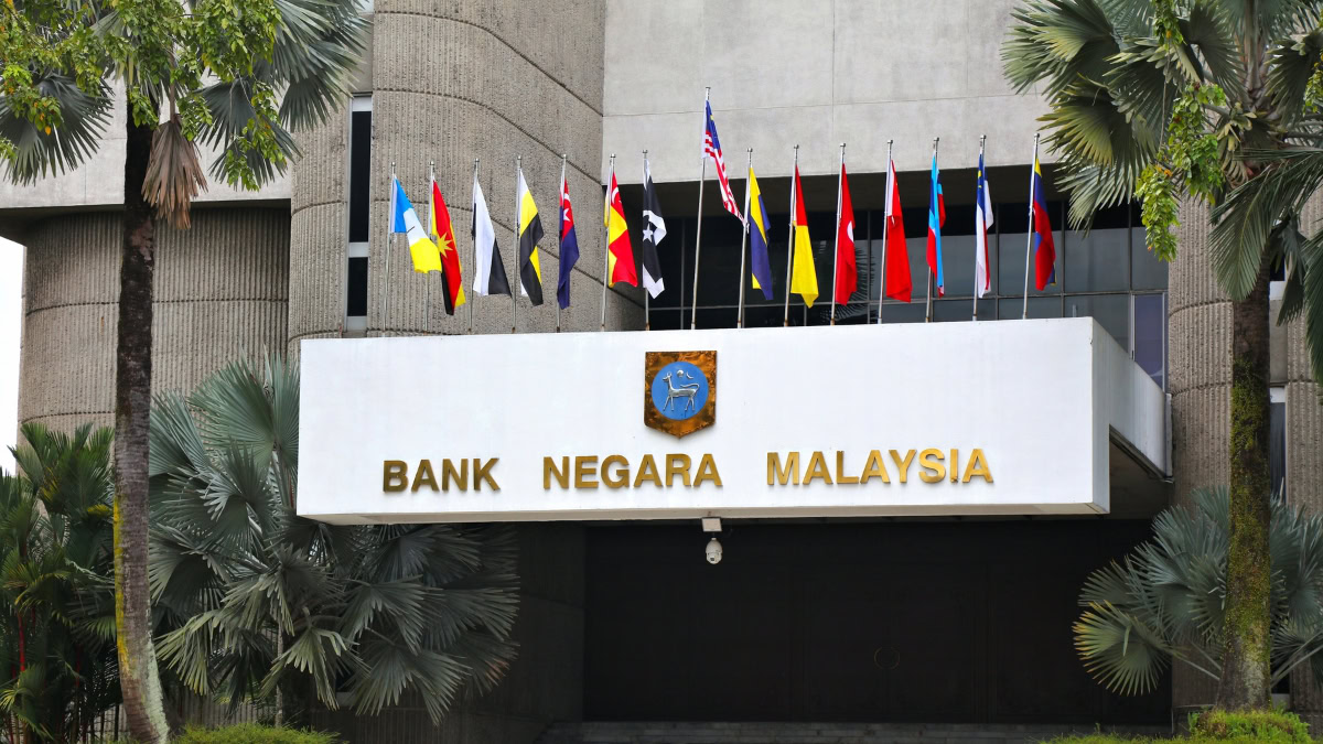 Front of Bank Negara Malaysia building with a row of international flags and palm trees on either side.