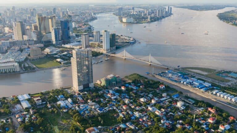 Aerial view of a modern city along a wide river, with a cable-stayed bridge, tall skyscrapers, and a mix of residential blocks nearby.
