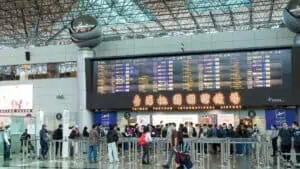 Busy airport check-in area with a large electronic flight information board above and crowds queuing at counters, Taiwan Taoyuan International Airport signage visible.